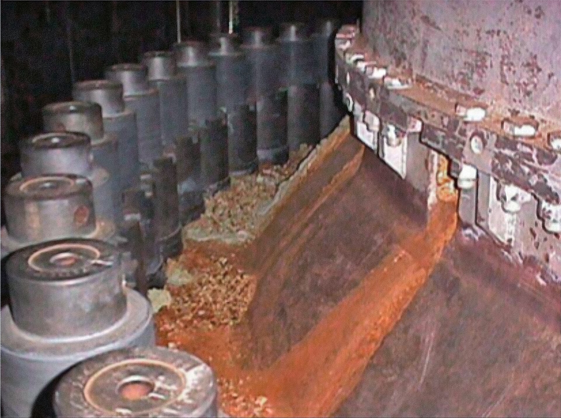 Orange rust-colored deposits piled against the steel surface of a pressure vessel head, with a row of grey control rod drive nozzles on the left