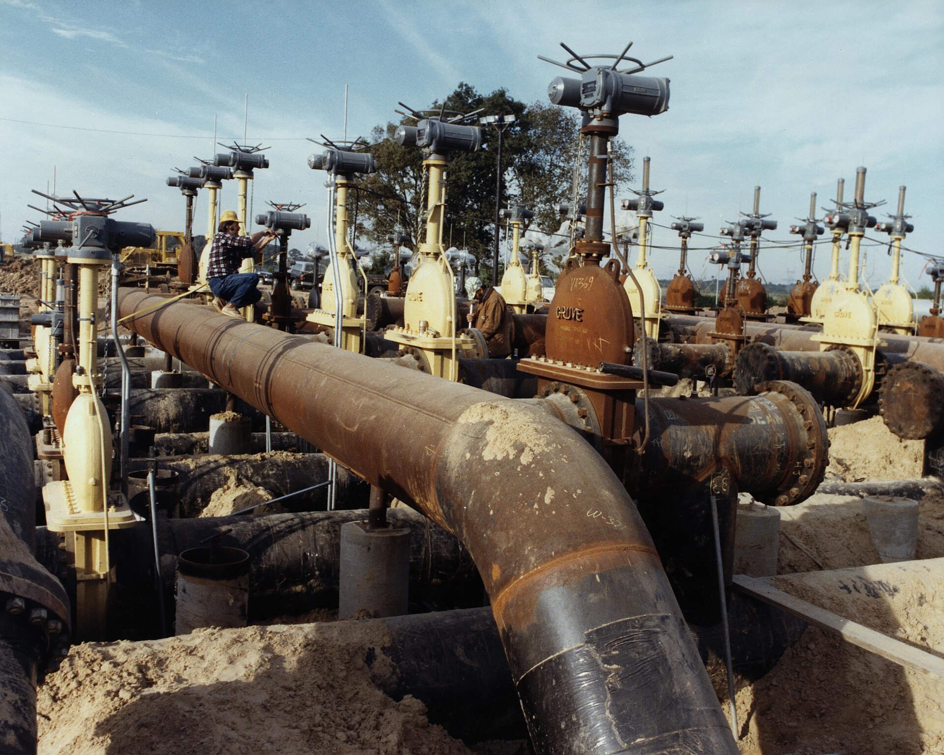Large-bore refinery piping with flanged gate valves and yellow electric actuators under construction, two workers at the valve stems