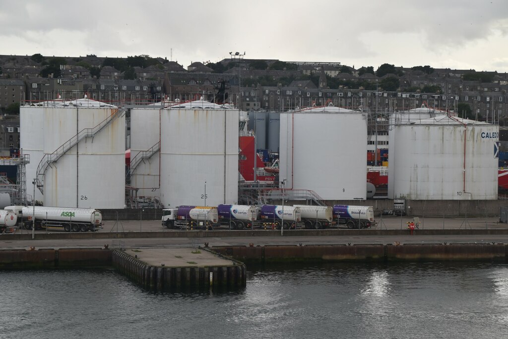 Numbered white aboveground storage tanks with external stairways at a harbour terminal, road tankers parked in front