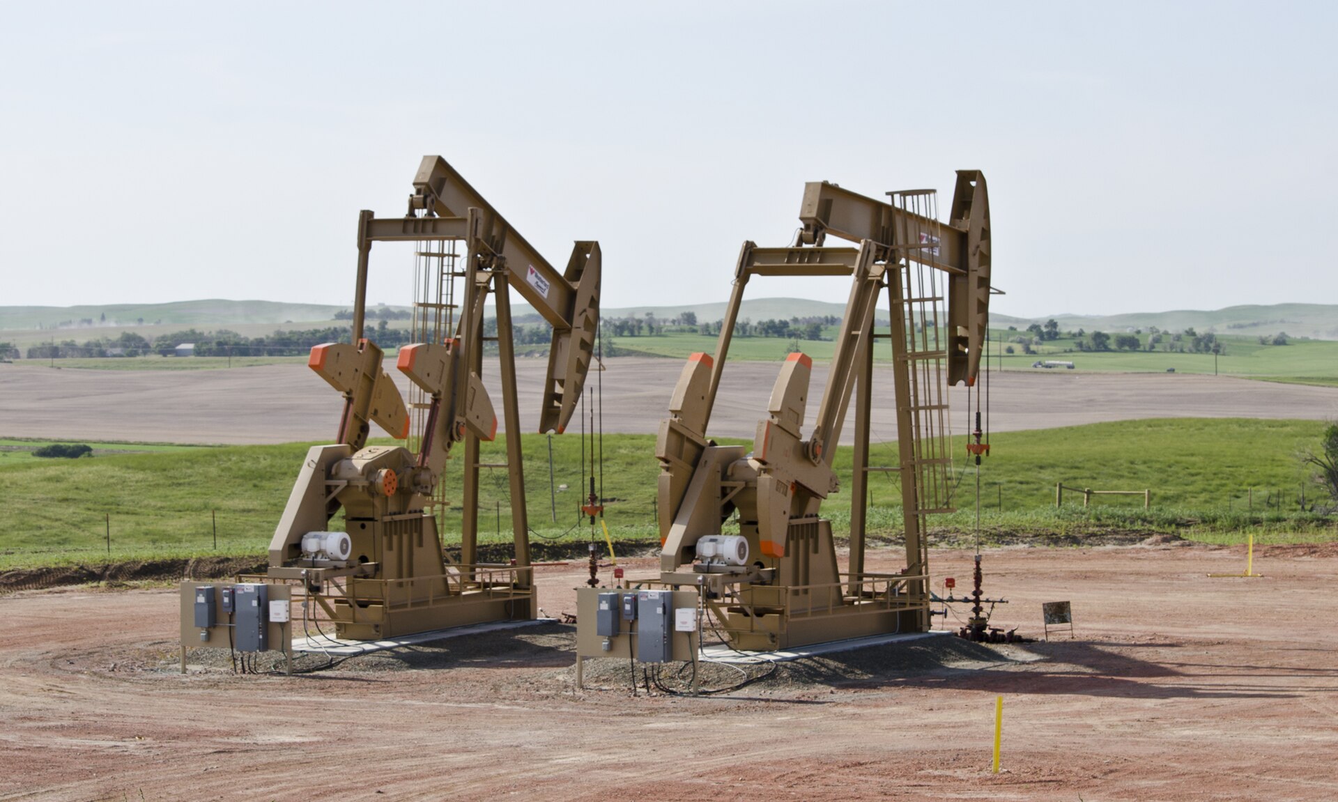 Two beam pumping units on a gravel well pad with farmland and low hills behind