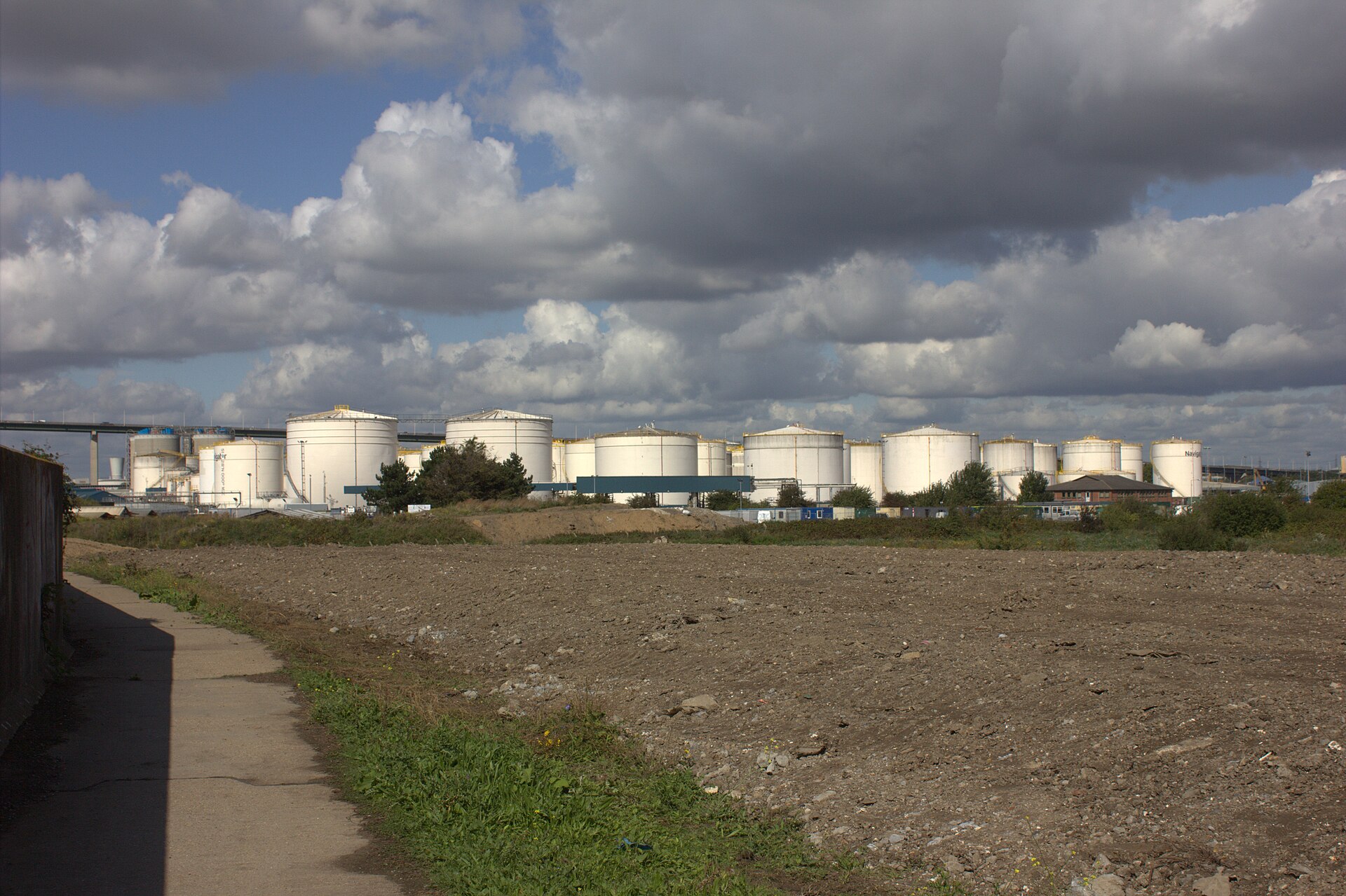 A row of large white oil storage tanks at a coastal tank farm under a cloudy sky