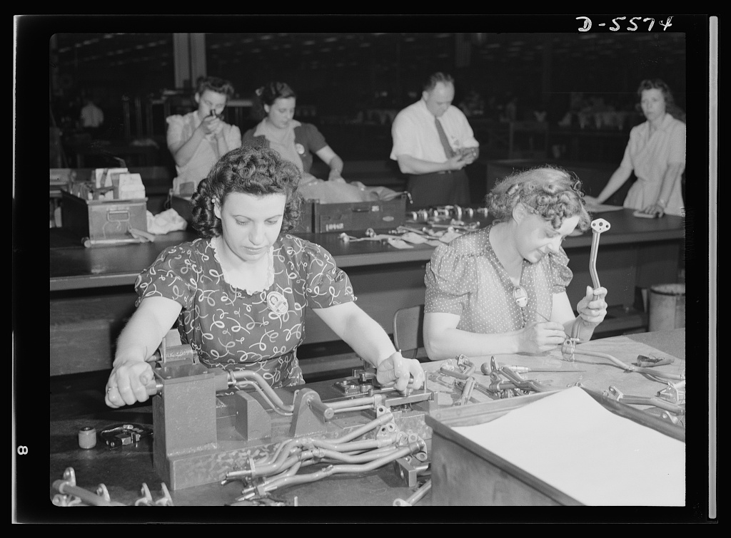 A woman inspector at a wartime factory bench checking machined parts on the inspection line