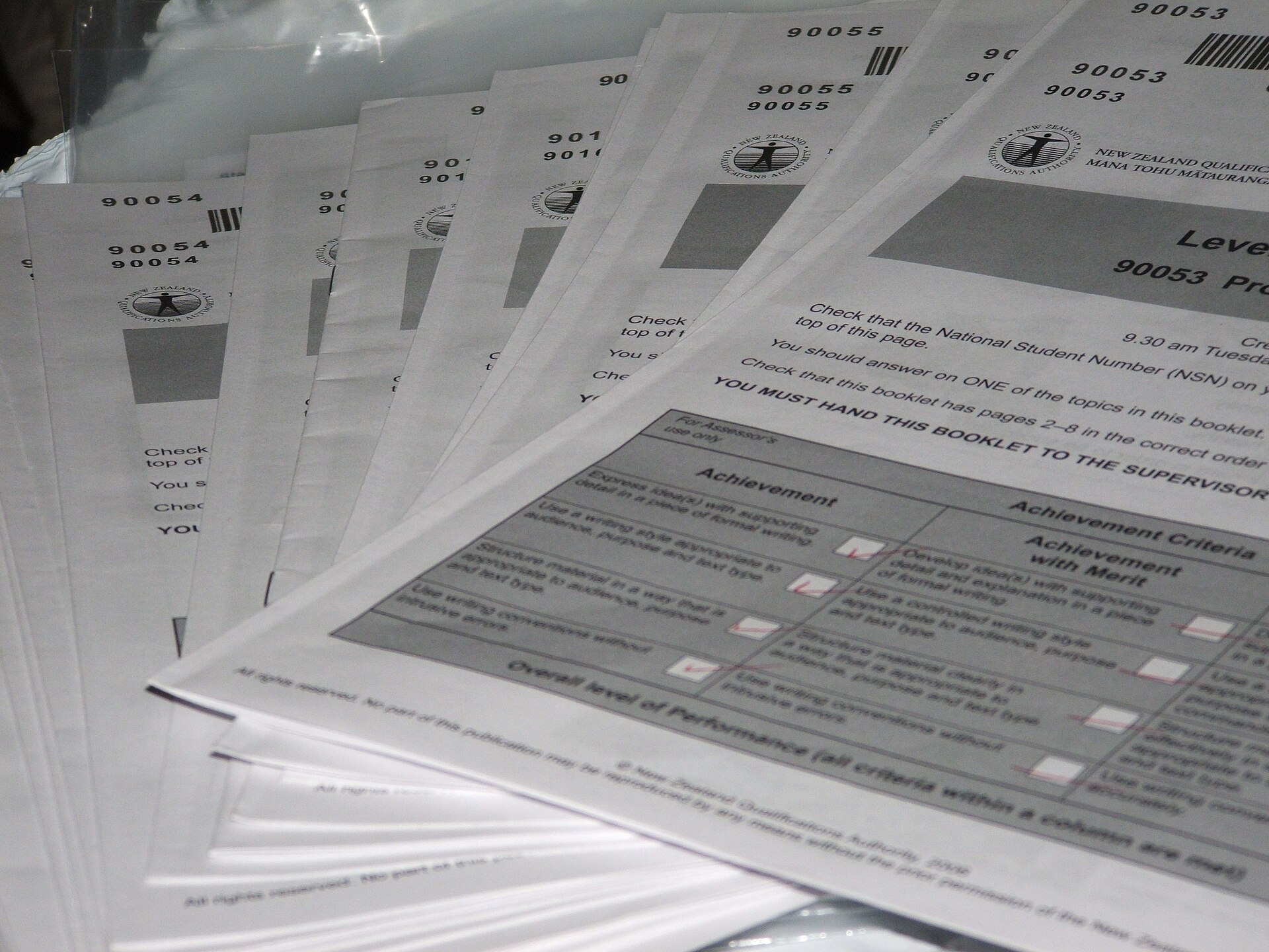 A fanned stack of printed exam booklets with assessment criteria tables on the cover