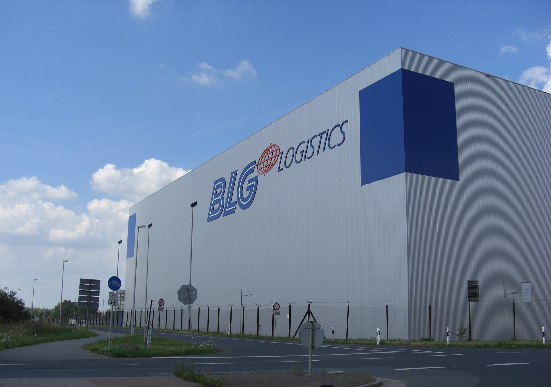 The tall grey side wall of a large logistics building under a blue sky, with a company logo near the roof edge and a security fence along the road