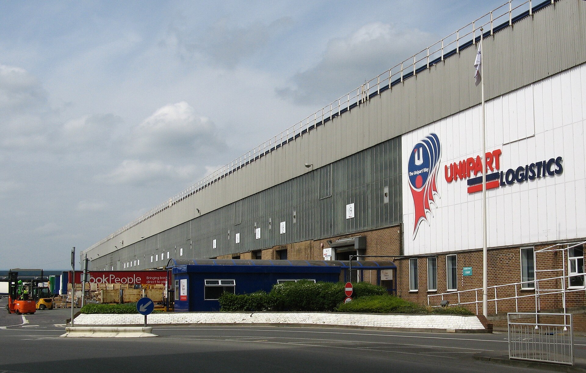The long loading-dock frontage of a logistics warehouse with numbered bay doors, a forklift on the left and a company sign on the white end wall