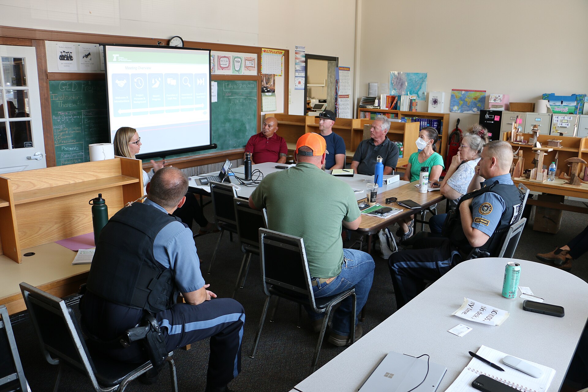 A project management team meeting around tables in front of a projected meeting overview slide